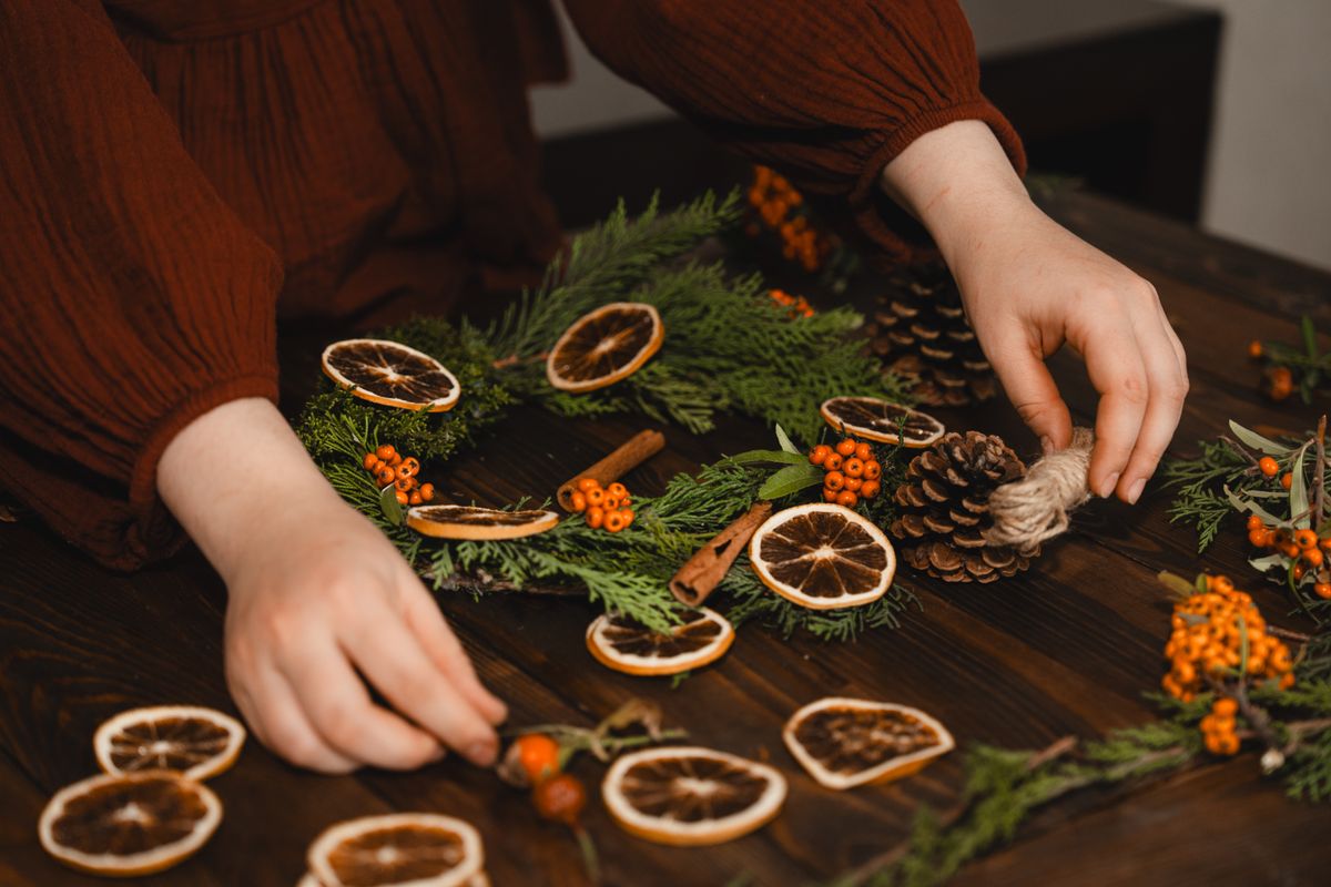 Hands assembling an evergreen wreath with dried oranges and pinecones on a dark wood table
